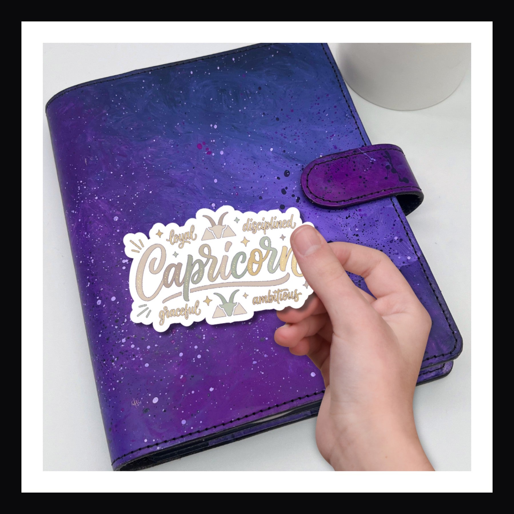 Hand holding a "Capricorn" sticker over a purple planner on a white background.