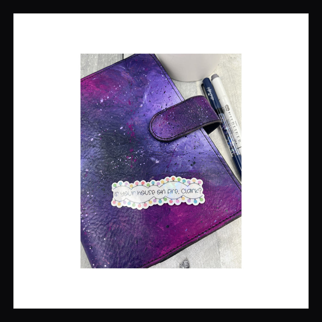Holographic die cut featuring 2 strands of multicolored christmas lights and the quote "Is your house on fire, Clark?"