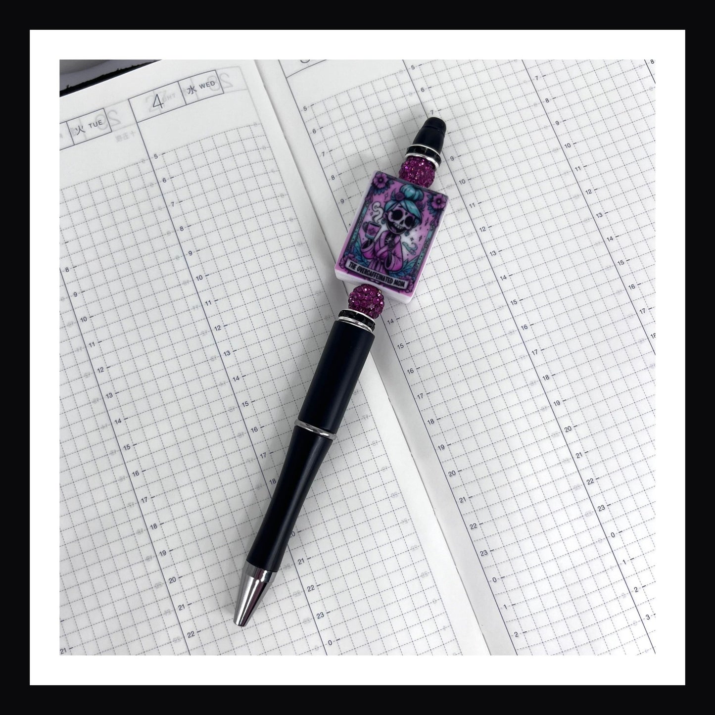 Over-caffeinated Mom Pen is a black ink ballpoint pen featuring a black barrel with two pink rhinestone beads and a large silicone bead with a printed tarot card style image of a skeletal woman with a cup of coffee.