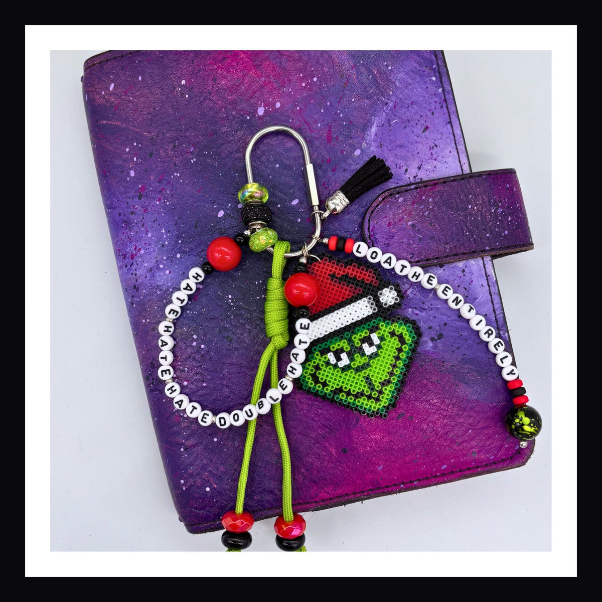 Loathe Entirely Carabiner Bag Charm features a handmade Grinch charm, a paracord knot with beads and two friendship-bracelet-style beaded tags that collectively read "Hate, hate, hate. Double hate. Loathe entirely".
