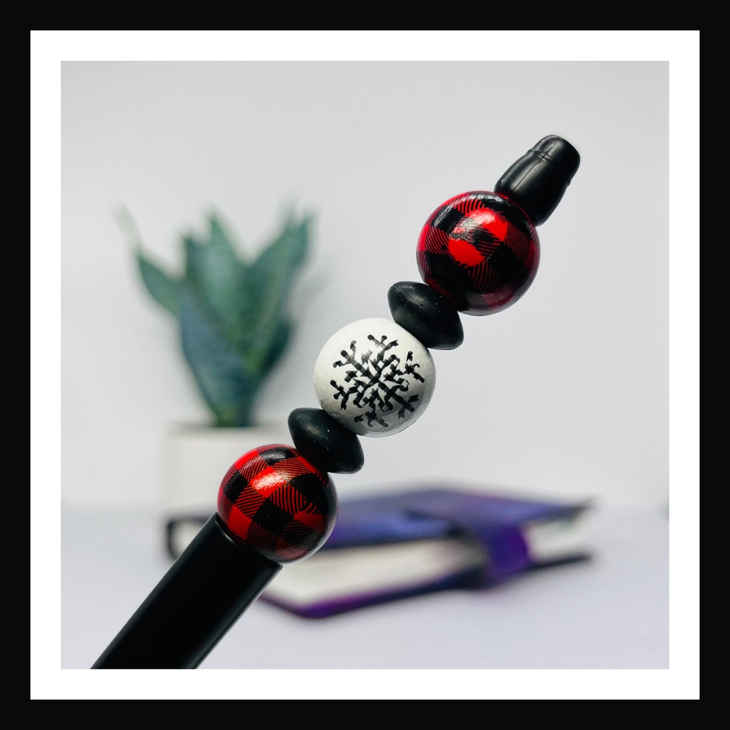 Snowflakes & Scarves Pen is a black ink ballpoint pen featuring 3 wood beads with two silicone spacers.  Two of the three beads feature red buffalo plaid, the third bead features a snowflake.