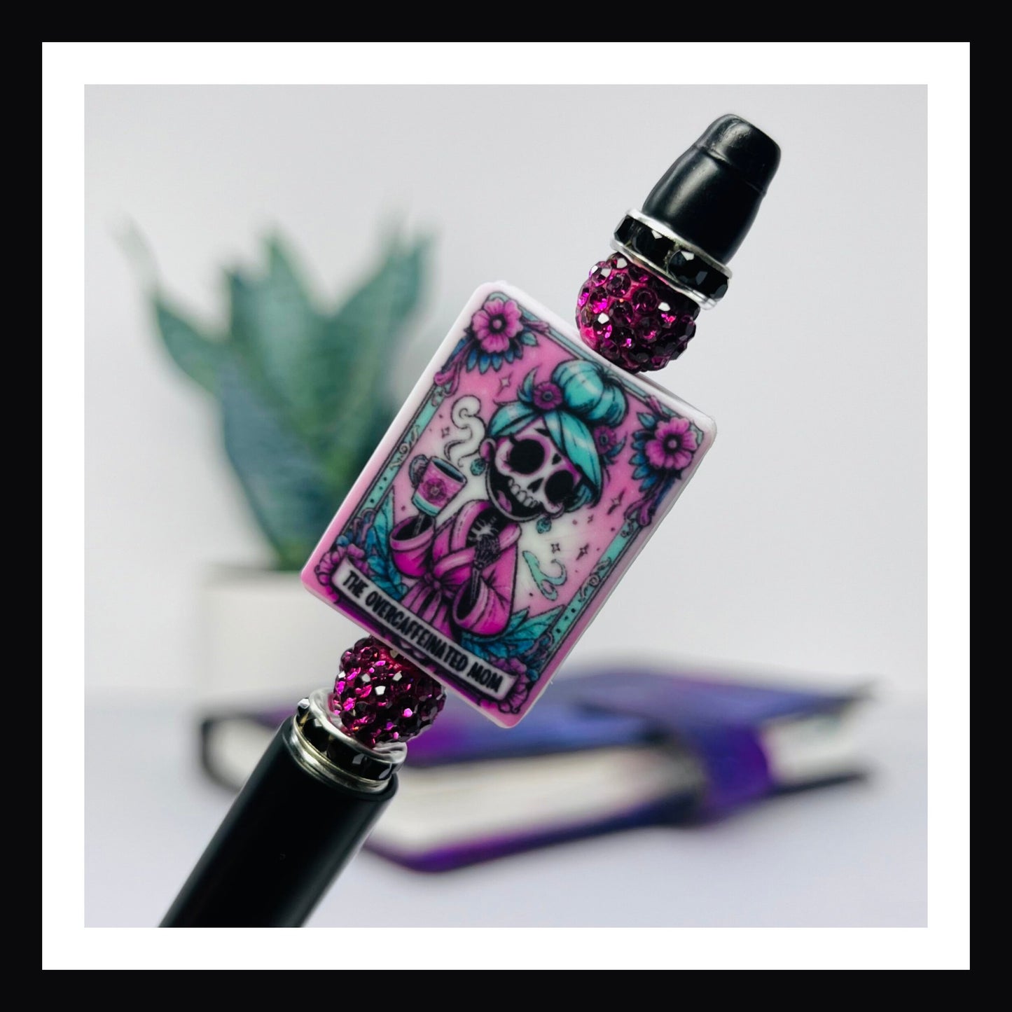 Over-caffeinated Mom Pen is a black ink ballpoint pen featuring a black barrel with two pink rhinestone beads and a large silicone bead with a printed tarot card style image of a skeletal woman with a cup of coffee.