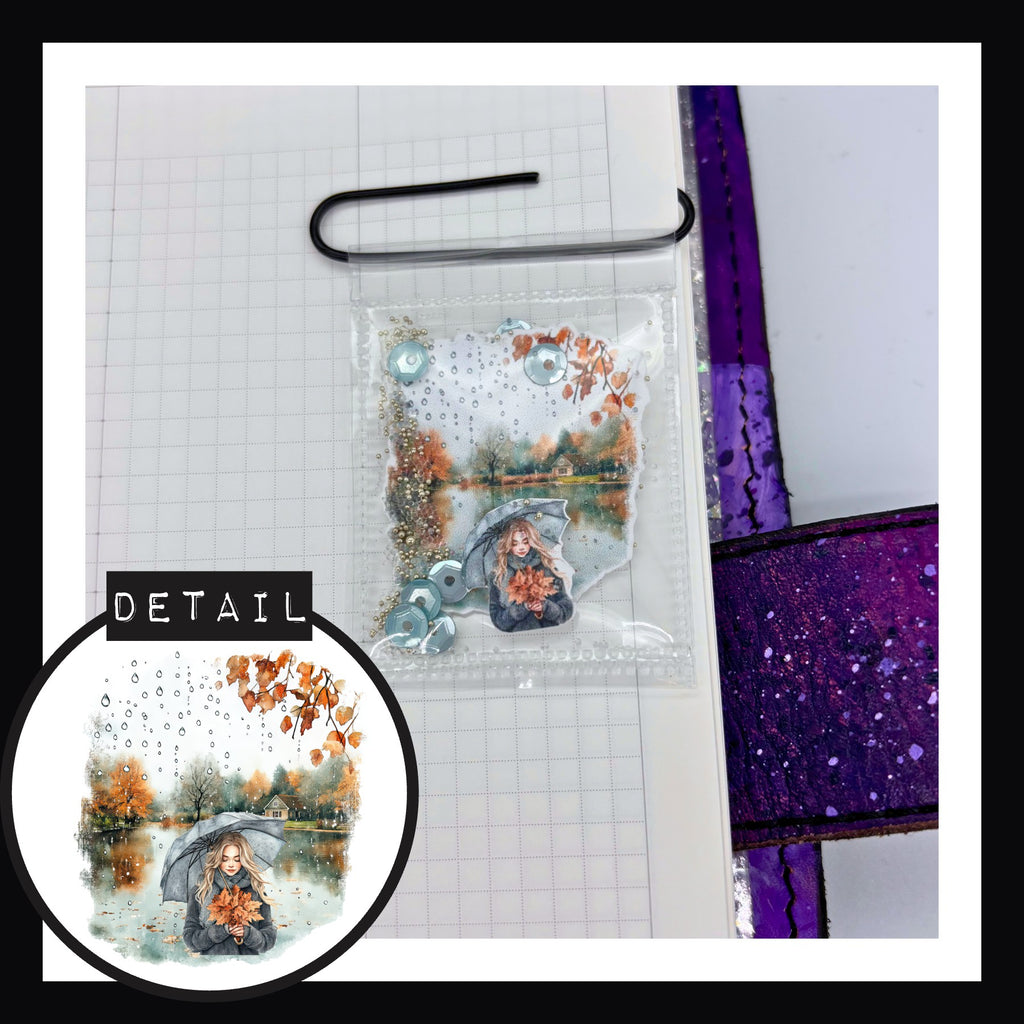 November Rain Shaker Paperclips Lakefront view features a design of a woman holding a bundle of leaves under umbrellas in front of a misty lake.  Shaker includes blue sequins, crystal and white gold caviar beads.