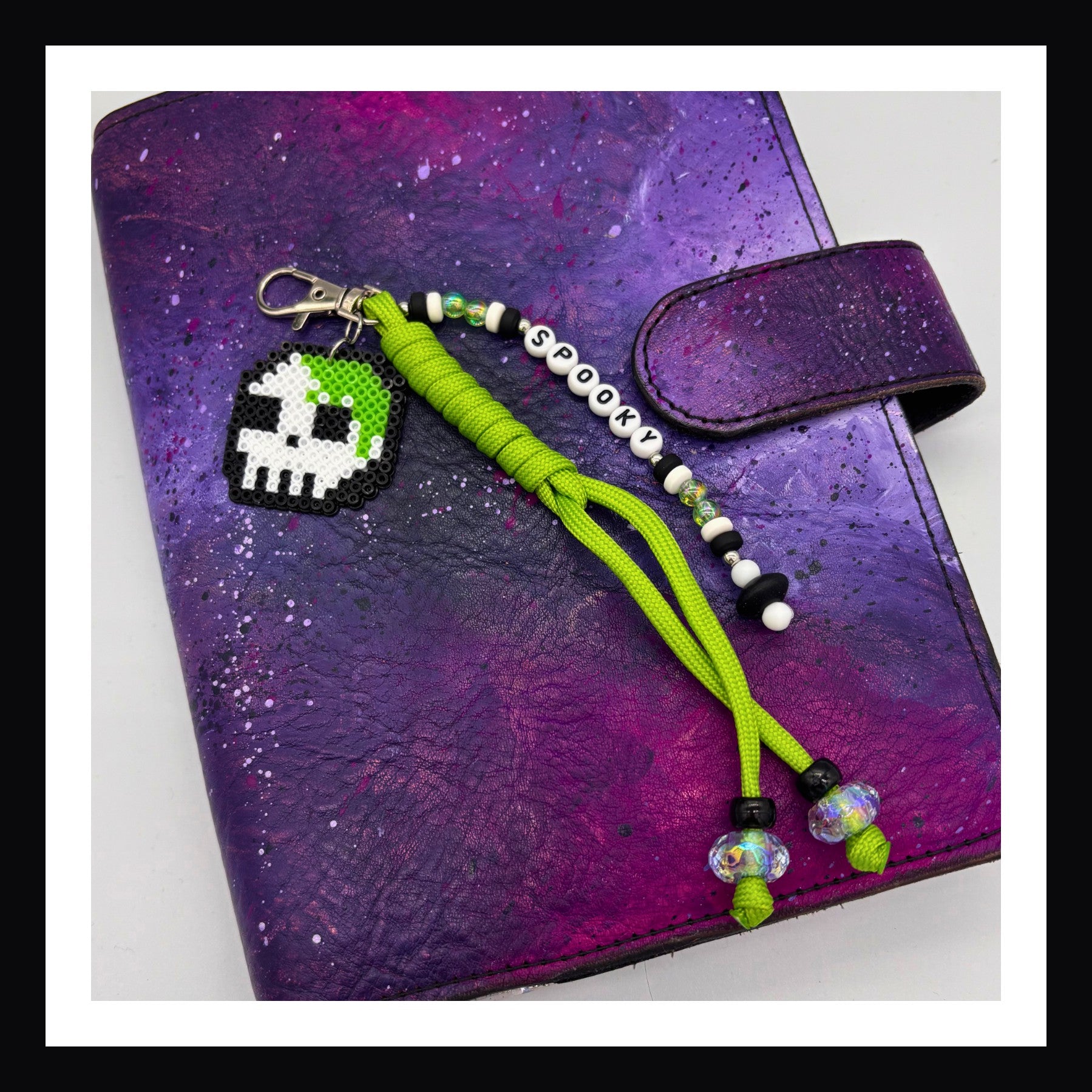 The Spooky Skull charm features a mossy green and white skull made of fuse beads with a matching braided paracord tassel and friendship bracelet style beaded tag that spells out "spooky" secured with a lobster clasp. 