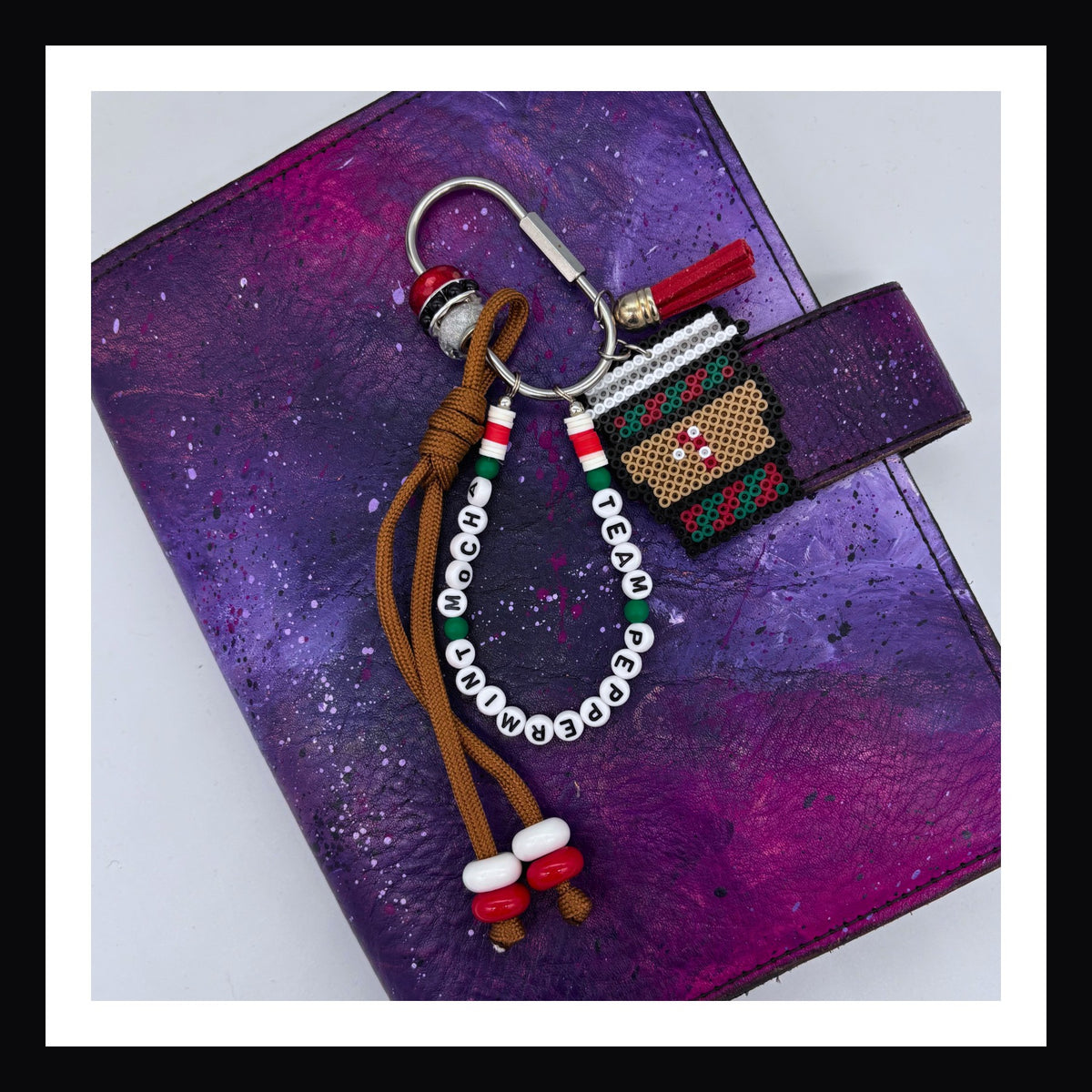 Peppermint Mocha Carabiner Bag charm features a handmade coffee cup charm with peppermint detail, a friendship-bracelet-style beaded tag that reads "team peppermint mocha", a knotted paracord with beaded detail and vegan leather tassel all on a silver carabiner with beads for additional sparkle.