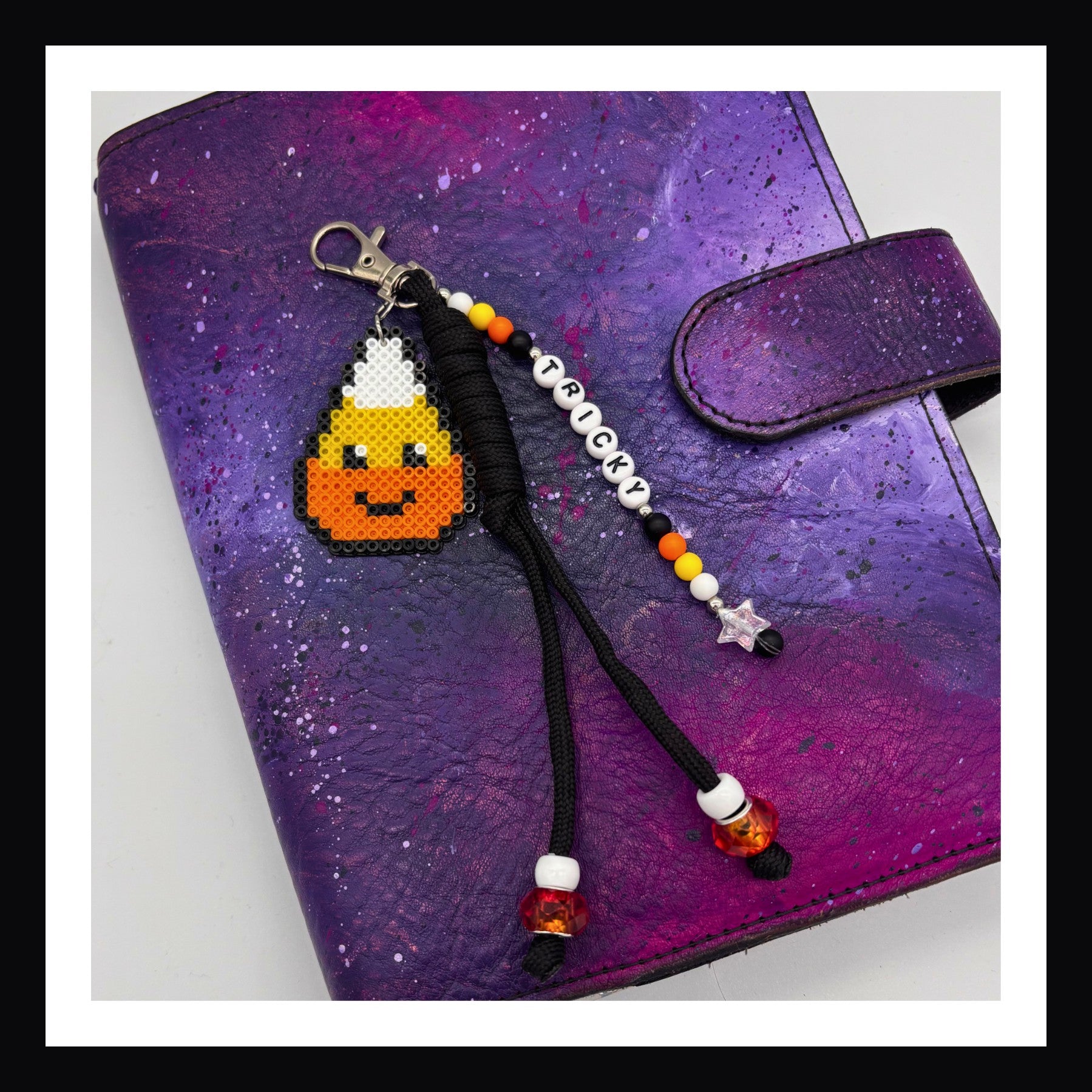 A candy corn charm with smiling face paired with black braided paracord and friendship bracelet style tag that spells out "Tricky".  Secure with a lobster clasp.